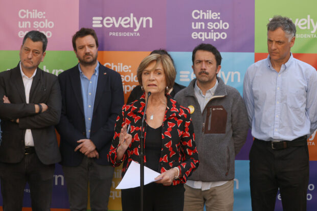 Evelyn Matthei habla de pie frente a un micrófono. Detrás de ella 4 integrantes de su equipos. De fondo una gigantografia colorida con consignas de su campaña.