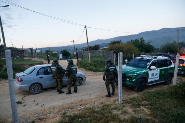 Tres carabineros de Chile con equipo táctico de pie en un camino de tierra junto a un vehículo civil y una patrulla verde durante un operativo en el asentamiento informal de Calicheros.