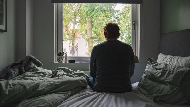 Un hombre de espaldas sentado en su cama ve árboles a través de la ventana.