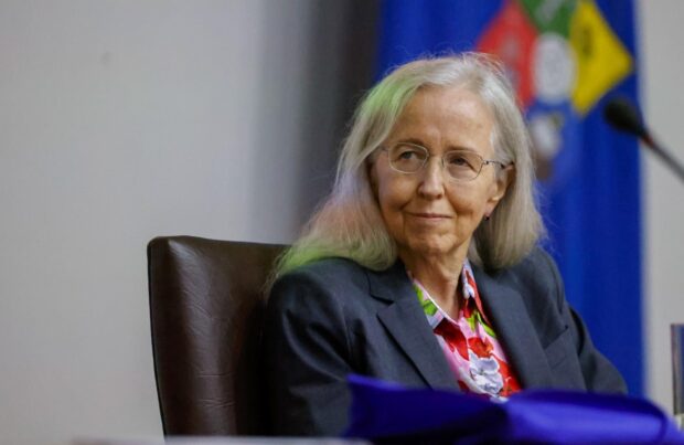 Elizabeth Horan sentada en una silla negra mirando hacia su izquierda. Atrás de ella se ve borrosa la bandera oficial de la Universidad de Chile.