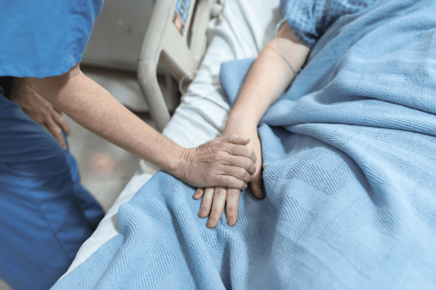 Una persona con uniforme médico o de cuidador sostiene y acaricia la mano de un paciente, que está acostado en una cama de hospital bajo una manta azul.