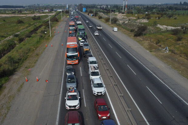 Vista aérea de una carretera en Chile que muestra un intenso flujo vehicular y congestión en una de las pistas. Se ven vehículos, incluyendo autos, camiones y un bus de pasajeros, detenidos o avanzando lentamente. La imagen capta el tráfico en un día soleado, con conos de seguridad en el borde de la vía.