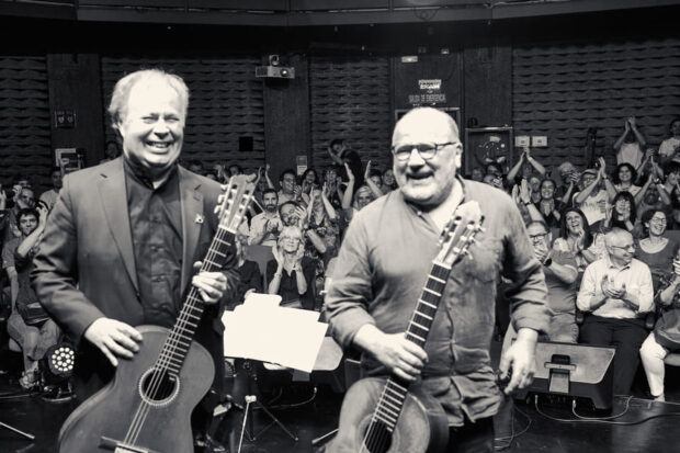 Imagen en blanco y negro de Horacio Salinas y Romilio Orellana sonriendo en un escenario con sus guitarras en mano. Detrás de ellos una multitud dentro de una sala.