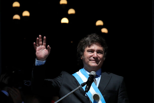 Javier Milei usando la cinta presidencial celeste y blanca de Argentina. Delante de un micrófono se ve saludando con una mano.