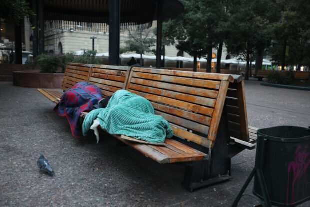 Personas en situación de calle durmiendo en una banca de madera arropadas con frazadas.