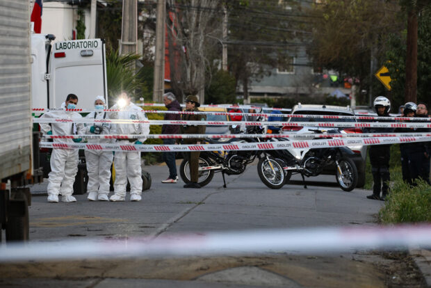 Investigadores forenses y carabineros vestidos con overoles blancos en una escena de crimen en una calle acordonada con cintas de seguridad.
