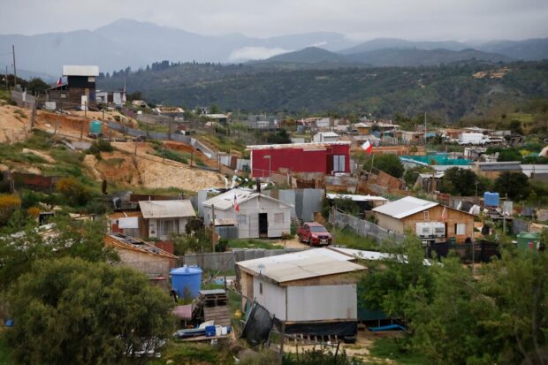 Vista panorámica del extenso asentamiento informal Calicheros en la Región de Valparaíso, con numerosas viviendas precarias de diferentes materiales dispersas en una colina árida y montañas nubladas de fondo.