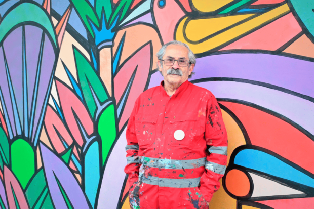 El muralista Mono González posando con un overol rojo frente a uno de sus murales.