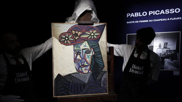 Dos hombres con delantales sostienen un retrato colorido y cubista de una mujer, una pintura de Picasso titulada 'Buste de femme au chapeau à fleurs'.