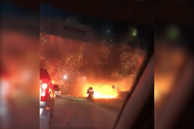 Toma oscura de una carretera vista desde el interior de un vehículo, donde una explosión de color naranja brillante con chispas ilumina la noche. Un vehículo con luces rojas está parado en el carril izquierdo en primer plano. Un par de vehículos están parcialmente visibles en el carril izquierdo y en el arcén. El fuego y el humo llenan el centro y la derecha de la escena.