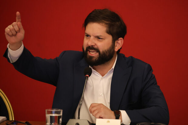 Presidente Gabriel Boric sentado, hablando y gesticulando con las manos a través de un micrófono en un salón con paredes rojas.