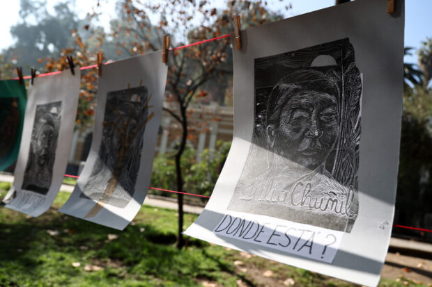 Cuatro grabados en blanco y negro con la imagen de Julia Chuñil, colgados de una cuerda con pinzas de ropa en un parque soleado. El grabado frontal lleva el texto 'Julia Chuñil' y 'DONDE ESTA?'.