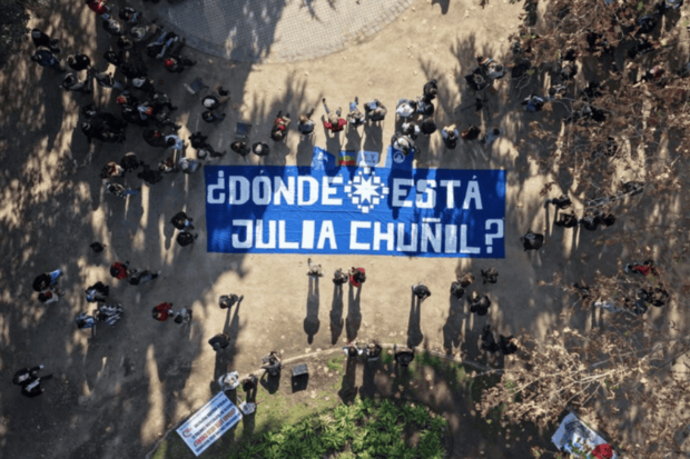 Vista aérea de una manifestación en un parque, donde un grupo de personas sostiene una gran pancarta azul con letras blancas que dice: '¿DÓNDE ESTÁ JULIA CHUÑIL?'"