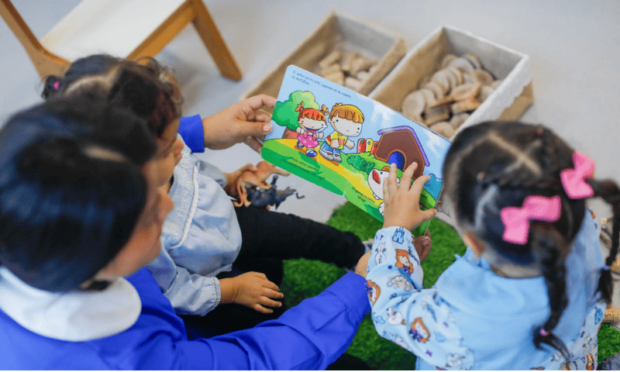 Una educadora de párvulos lee un libro ilustrado a tres niñas en el suelo, rodeadas de materiales didácticos de madera. La imagen representa la educación inicial y el fomento de la lectura.