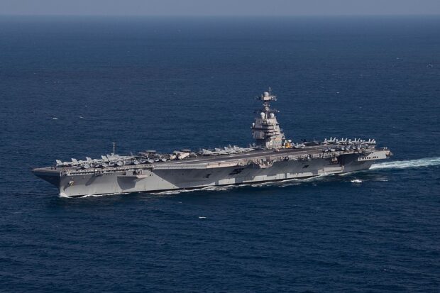 Vista aérea lateral del portaaviones USS Gerald R. Ford (CVN-78), navegando en aguas profundas de color azul oscuro. El gran buque de guerra gris tiene aviones de combate y helicópteros estacionados en su cubierta de vuelo.