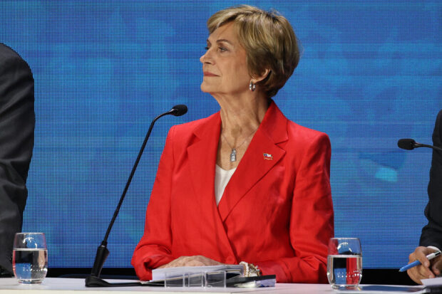  Retrato medio de Evelyn Matthei, con el cabello rubio corto, mirando hacia la izquierda mientras está sentada en una mesa durante el evento ENADE. Viste un blazer rojo sobre una blusa blanca, un collar con un dije y un pin con la bandera de Chile en la solapa. Detrás de ella hay un fondo azul texturizado.