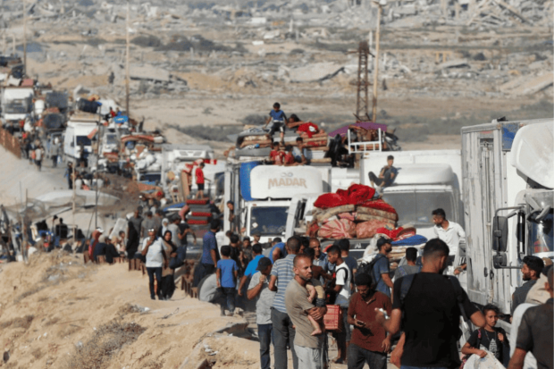 Larga fila de personas desplazadas caminando junto a camiones y vehículos sobrecargados de pertenencias en un camino de tierra, con ruinas de edificios destruidos en el horizonte.