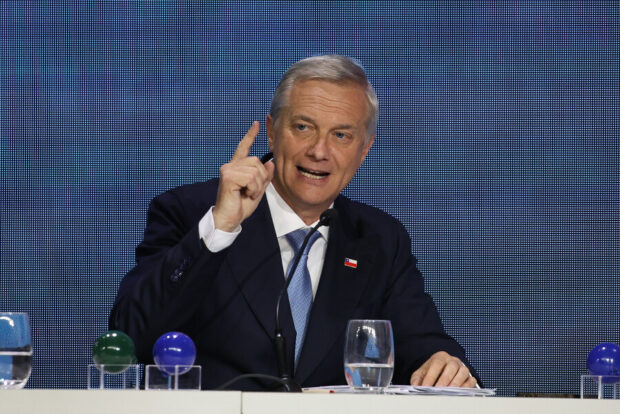 José Antonio Kast sentado en una mesa durante el evento ENADE. Viste un traje azul marino con una corbata azul y una camisa blanca, y lleva un pin con la bandera de Chile en la solapa. Su mano derecha está levantada con el dedo índice señalando hacia arriba, gesticulando mientras habla.