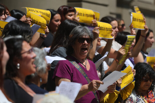  Un grupo de mujeres protestando, con una mujer mayor en primer plano que lleva gafas de sol, una camiseta morada, y sostiene papeles mientras grita con pasión. Varias otras mujeres detrás de ella sostienen carteles de color amarillo brillante que dicen "basta de impunidad".