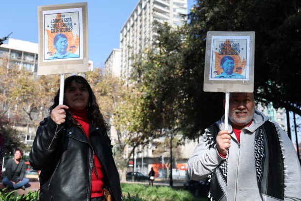 Dos manifestantes, un hombre y una mujer, sostienen carteles idénticos preguntando por Julia Chuñil Catricura. Los carteles tienen el rostro de la mujer y el texto "¿DÓNDE ESTÁ JULIA CHUÑIL CATRICURA?". La escena es en un parque urbano,