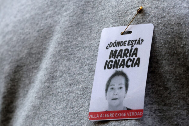 Pin o identificación con la foto de una mujer y la pregunta '¿DÓNDE ESTÁ? MARÍA IGNACIA', sujeto a una tela gris con un alfiler de gancho.