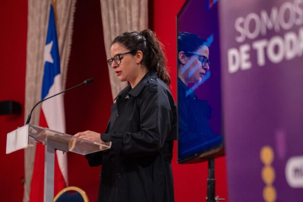 Ministra Orellana habla desde un podio en un salón con paredes rojas. A su lado se ve la bandera de Chile.