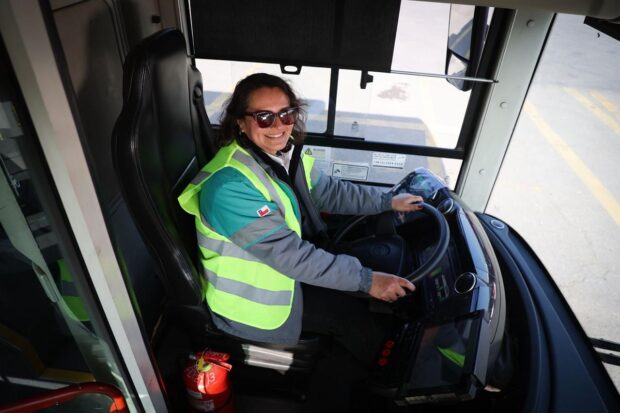Una mujer sonriente, con lentes de sol, está sentada en el asiento del conductor de un autobús. Viste una chaqueta con mangas grises y un chaleco reflectante de color amarillo neón y turquesa. Ambas manos están sujetando el volante.
