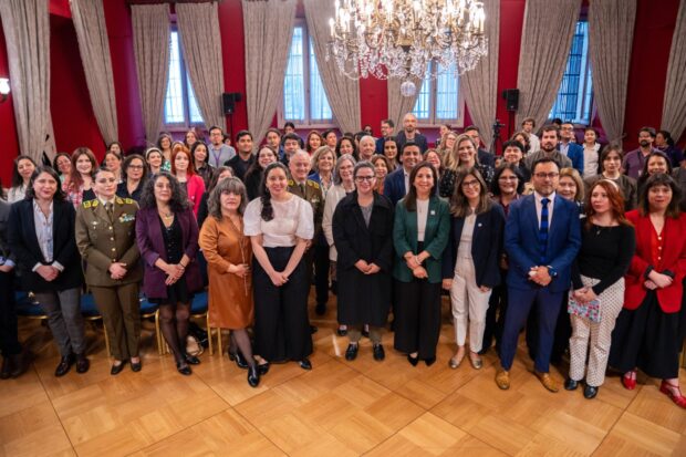 Foto grupal de aproximadamente 40 personas, mayoritariamente mujeres, incluyendo autoridades civiles y una militar uniformada, posando frente a una sala de conferencias con paredes rojas, grandes cortinas y un candelabro.