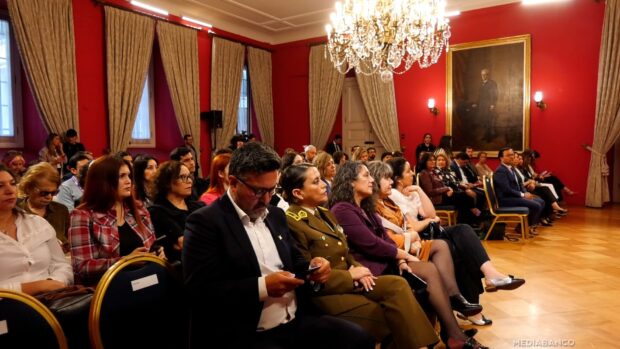 Asistentes sentados, incluyendo civiles y una mujer uniformada, en la presentación del estudio sobre Violencia Política Digital de Género, en una sala con paredes rojas, grandes cortinas y un candelabro.