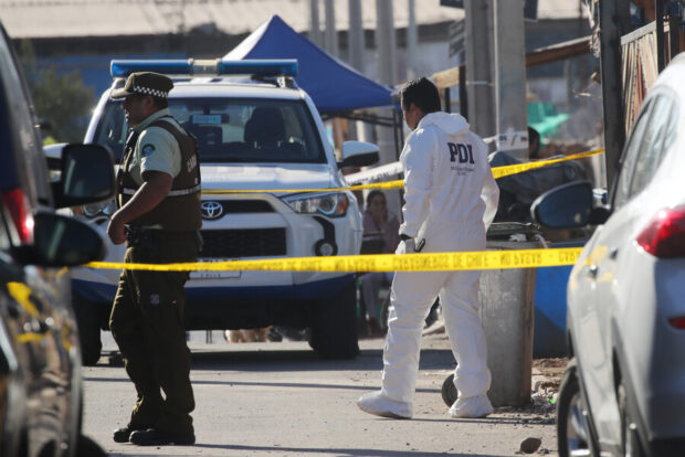 Funcionario forense de PDI vestido con overol blanco mientras realiza trabajo pericial detrás de una cinta de seguridad amarilla. Se observa una camioneta policial blanca con una baliza azul a la izquierda y una carpa azul al fondo.