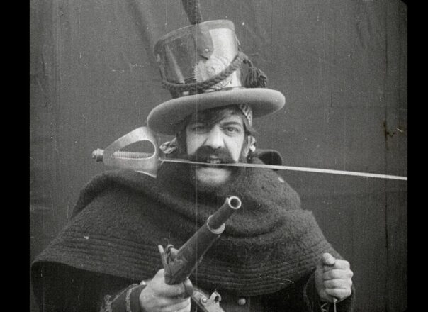 Primer plano en blanco y negro de un hombre con un bigote grueso y un sombrero de húsar, sosteniendo una pistola de chispa en primer plano y mordiendo una espada con la boca, con la punta de la hoja sobresaliendo por detrás de su cabeza. El hombre lleva una capa oscura; la escena es un encuadre dramático de la película El Húsar de la Muerte.