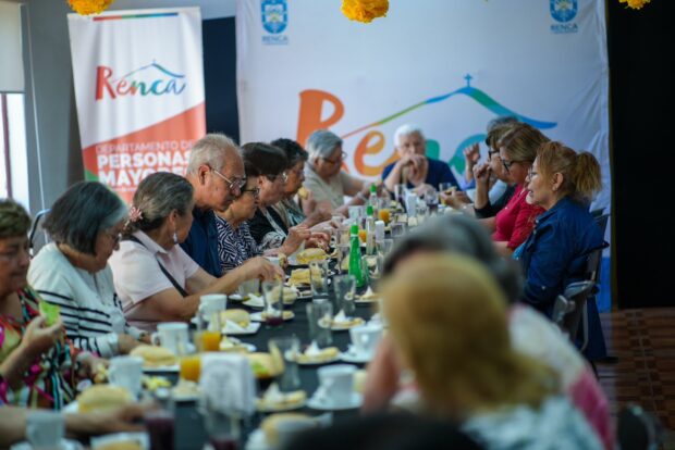 Una toma de ángulo ligeramente bajo de un grupo de adultos mayores sentados alrededor de una mesa larga y oscura, compartiendo un desayuno. La mesa está llena de tazas, platos con sándwiches y jugos.