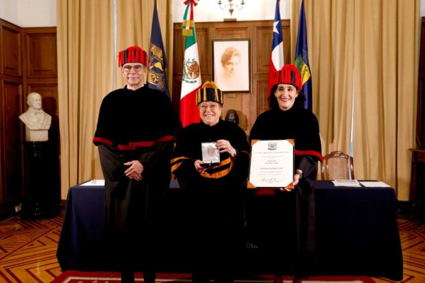 Michelle Bachelet posa de pie sosteniendo una medalla ceremonial entre dos autoridades académicas de la UNAM. Los tres visten togas negras con bandas y birretes doctorales.