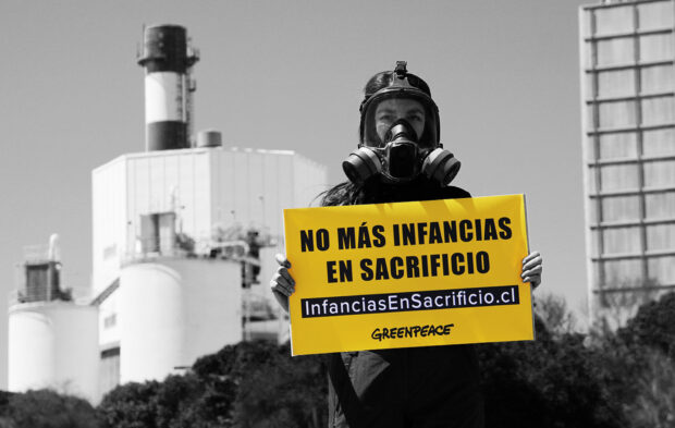 Fotografía en blanco y negro (con el cartel amarillo resaltado) de una activista de Greenpeace usando una máscara de gas y un casco, parada frente a una gran planta industrial con chimeneas y edificios de tanques. La activista sostiene un cartel amarillo con letras negras que dice: 'NO MÁS INFANCIAS EN SACRIFICIO' y la dirección 'InfanciasEnSacrificio.cl / GREENPEACE'.