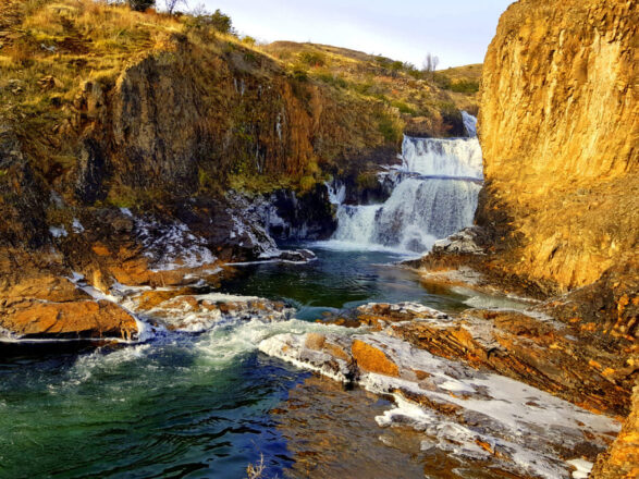Vista panorámica de la Cascada Los Maquis, donde el agua cae en varios niveles entre dos cañones rocosos de color dorado y ocre. El agua en la base es de color turquesa, y se observa algo de espuma en las rocas en primer plano
