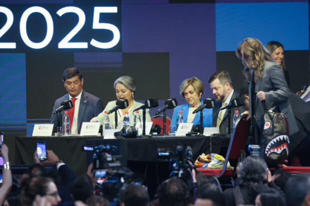 Candidatos presenciales chilenos sentados en una mesa de debate con micrófonos, frente a una audiencia y una pantalla de fondo que muestra el número "2025". Una mujer vestida de gris se retira por la derecha.