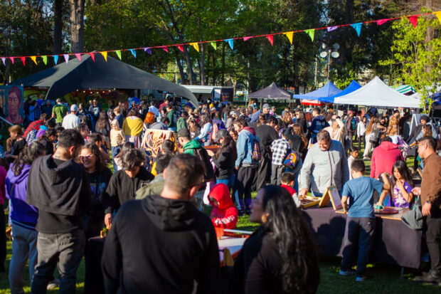 Gran multitud de personas reunidas en una feria o festival al aire libre en un parque. Hay puestos de venta bajo carpas, banderines de colores colgando y un ambiente animado con mucha gente.