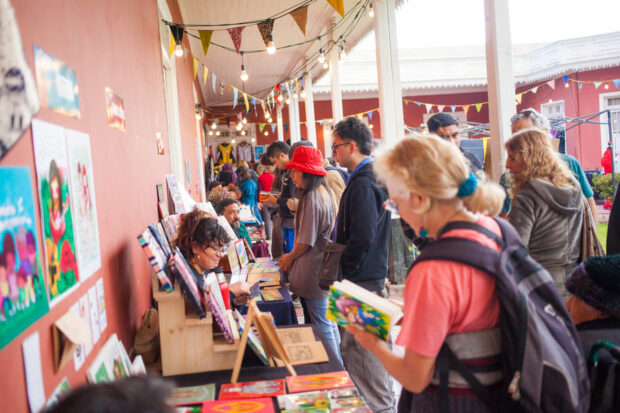 Fila de personas examinando libros y productos en estantes de una feria cultural al aire libre, con banderines decorativos y un techo con luces colgantes.