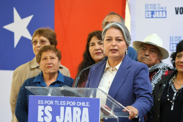 Jeannette Jara habla desde un podio con un cartel que dice: "Es por ti. Es por Chile. Es Jara". Detrás de ella ciudadanos adultos mayores y de fondo la bandera de Chile.