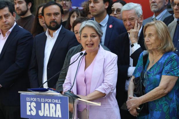 Jeannette Jara habla desde un atril transparente con el eslogan azul 'ES POR TI. ES POR CHILE. ES JARA'. Está rodeada por un grupo diverso de personas.
