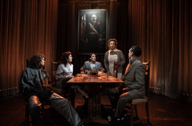 Escena de la obra La Fundación: Negacionismo y disputa de la verdad en Chile, con cinco actores y actrices sentados alrededor de una mesa bajo la mirada de un gran retrato militar, en un ambiente teatral iluminado con tonos cálidos.