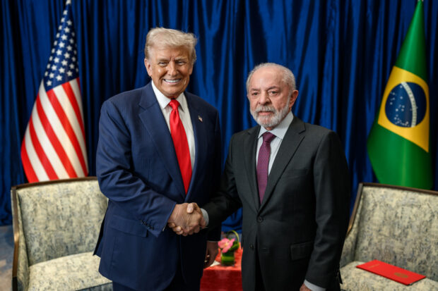 Donald Trump y Luiz Inácio Lula da Silva posan sonrientes mientras se estrechan de manos, atrás de ellos la bandera de Estados Unidos y la de Brasil.