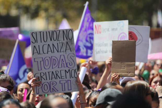 Primer plano de una gran multitud de mujeres en una manifestación del 8M bajo el sol. Varias manos levantan carteles escritos a mano. El cartel principal en primer plano dice en letras moradas: 'SI TU VOZ NO ALCANZA, TODAS GRITAREMOS POR TI'. En el fondo, se ven banderas moradas y otros mensajes de protesta.