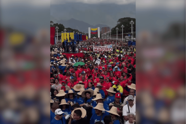 Gran multitud de personas, muchas vestidas con camisetas rojas y algunas con sombreros de paja, reunidas en una manifestación política masiva. Al fondo se distingue una bandera venezolana y un cartel con el texto "PSUV... CON MADURO'"