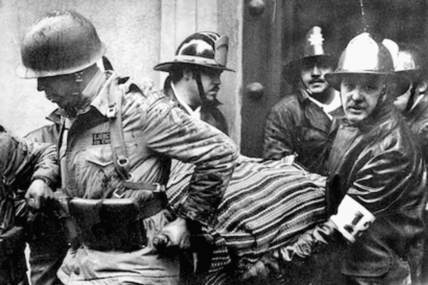 Fotografía en blanco y negro mostrando a un militar con casco y uniforme, junto a bomberos con cascos de estilo antiguo, cargando a una persona envuelta en una manta rayada durante un evento violento o de emergencia en un entorno urbano.