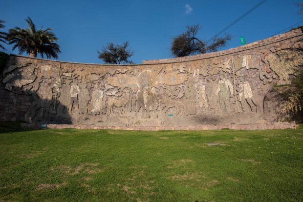Mural Tupahue en mosaico de piedra. El mural curvo representa figuras ancestrales y símbolos latinoamericanos. Se observa un tramo de césped en primer plano y árboles y cielo azul en la parte superior.