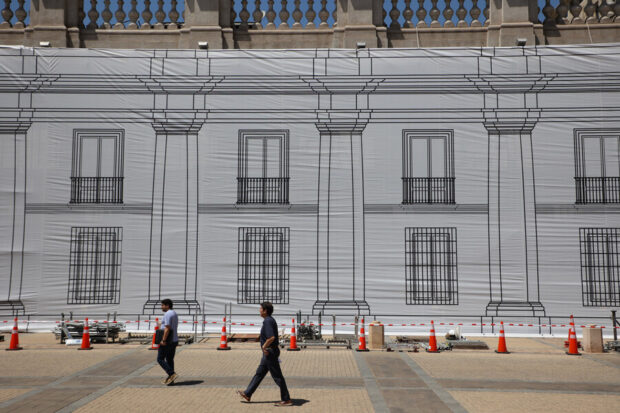 "Primer plano de una sección del Palacio de La Moneda en remodelación, cubierta por un gran lienzo blanco que tiene impreso un dibujo lineal en negro de la fachada arquitectónica con sus ventanas y balcones. Dos hombres caminan por el área peatonal frente a la lona, delimitada por conos de seguridad y vallas.