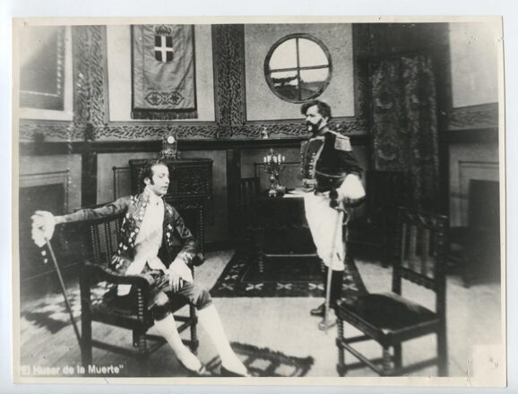 Fotografía en blanco y negro de la película muda chilena El Húsar de la Muerte (1925), que muestra una escena de interior. Un hombre elegantemente vestido con ropaje de la época colonial está sentado en una silla, y a su derecha, de pie y con uniforme militar de época, hay otro hombre con barba. Se observa una bandera y una ventana redonda en el fondo.
