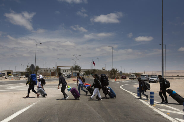 Familia de migrantes, incluyendo adultos y niños, cruzando a pie la frontera entre Chile y Perú, llevando grandes cantidades de equipaje en un día soleado.