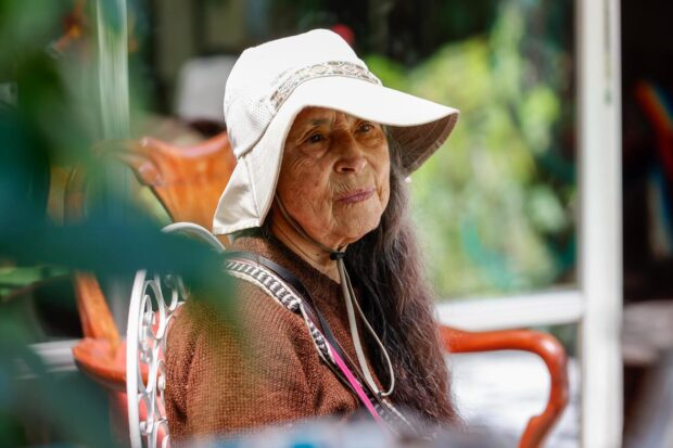 Mujer adulta mayor sentada al aire libre, con sombrero claro y vestimenta oscura, mirando hacia un costado, en un entorno natural y doméstico.